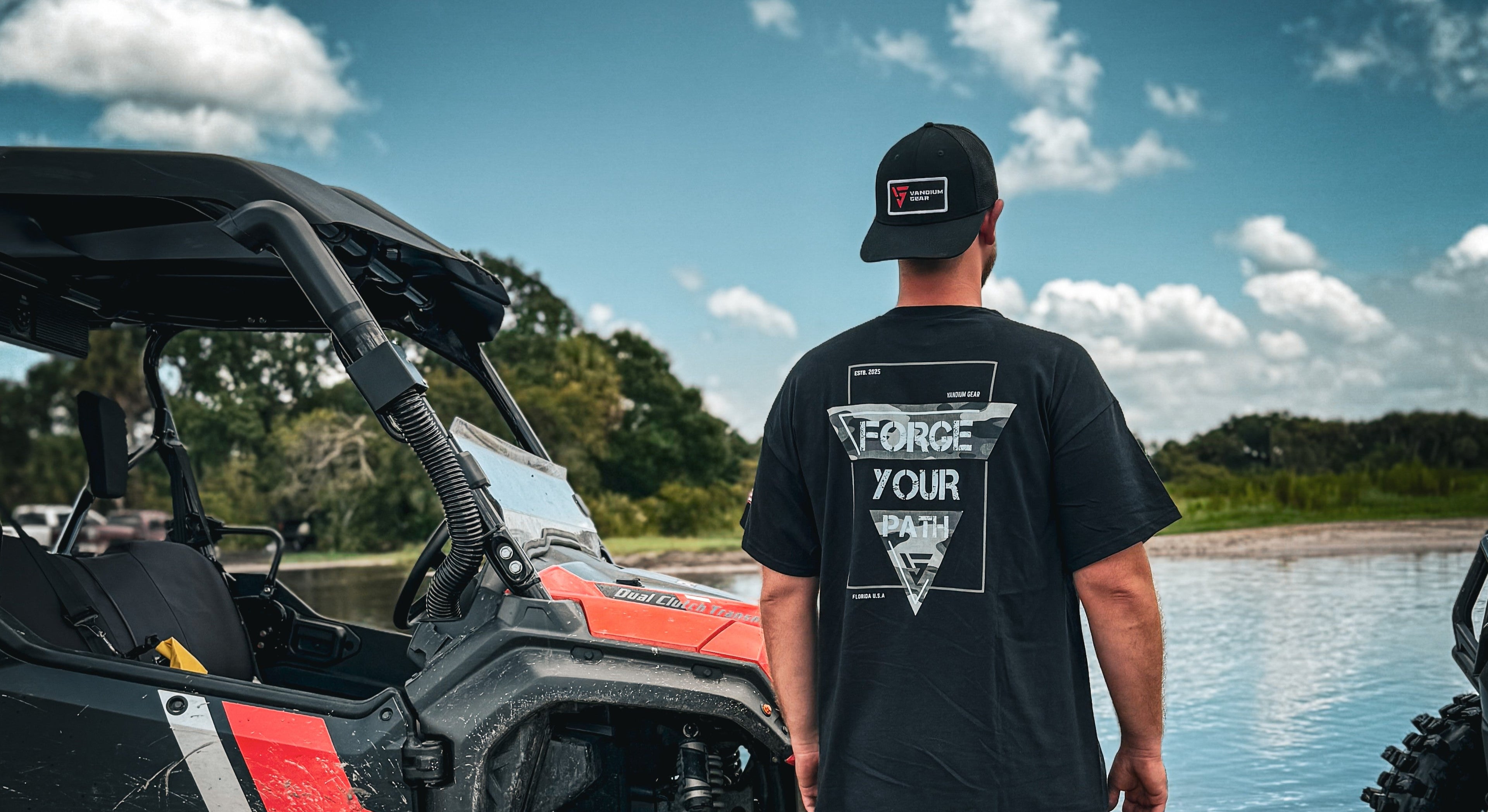 Person wearing a black t-shirt with 'Force Your World' design standing next to an off-road vehicle by a body of water.